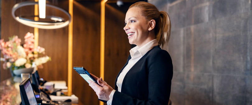 Hotel reception staff member holding a tablet behind a desk in a modern lobby with warm lighting and decorative elements